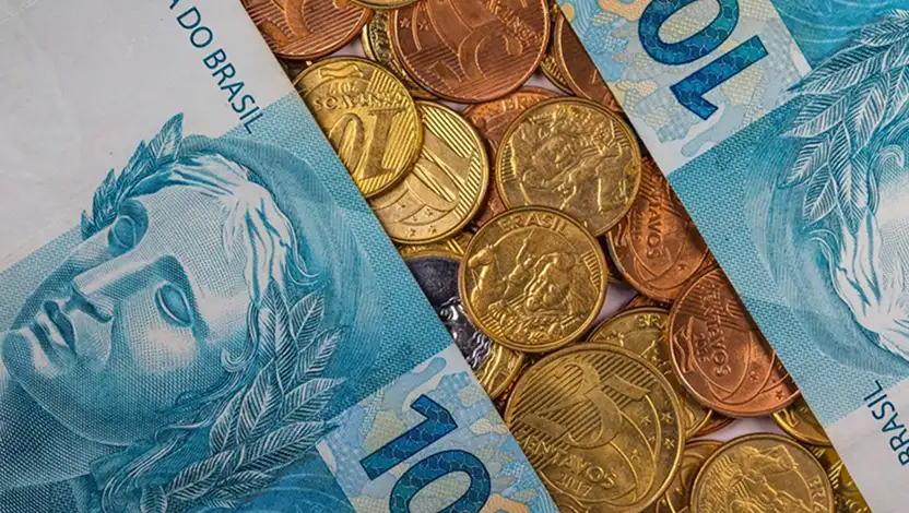 Moedas brasileiras e cédulas de 1, 5 e 10 reais sobrepostas, com destaque para notas azuis e moedas do real, sugerindo dinheiro e pagamento no Brasil.