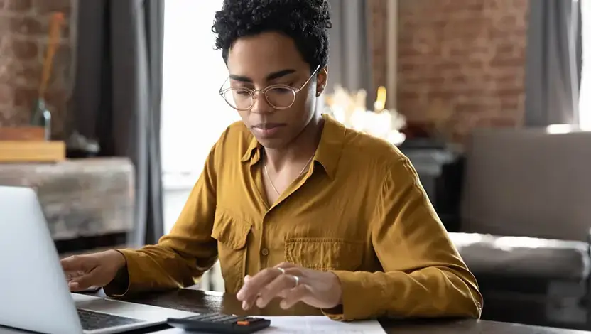 Pessoa com cabelo crespo usando óculos de grau trabalha em um laptop em ambiente de escritório, digitando e focada, com roupas casuais e luz suave ao fundo.