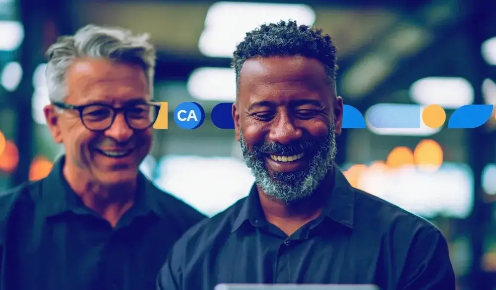 Dois homens sorrindo em frente a uma tela com fundo desfocado, transmitindo uma experiência de atendimento ou reunião corporativa em ambiente moderno.